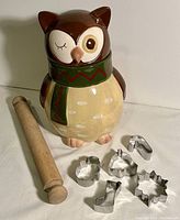 Photo shows vintage ceramic owl cookie jar with one eye winking, detailed green scarf with red zigzag pattern, beige belly with white spots, small feet, wooden rolling pin, and six metal holiday cookie cutters arranged on white surface.