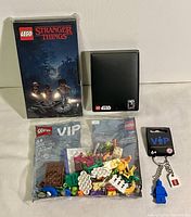 Photo showing all four Lego VIP goodies together: Stranger Things notebook with slipcover, Star Wars 40th Anniversary coin boxed, Spring Fun VIP Add-On Pack sealed in bag, and blue Lego mini-figure keychain in packaging.