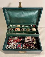 Photo of open green velvet lined jewelry box showing top compartment with various vintage costume jewelry pieces including costume earrings, brooches, and bracelets, with bottom compartment visible also filled with assorted jewelry.