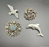 Four vintage silver toned brooches displayed on black background including bird, dolphin, floral wreath, and filigree floral with blue enamel