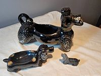 Photo showing all three items together on a table: black ceramic poodle-shaped serving dish with open space in center, ceramic dog ashtray/incense holder with gold accents lying down, small onyx carved dog figurine in front.