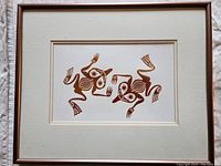 Full view of framed print depicting two stylized frogs in metallic ink, with a beige mat and wooden frame.