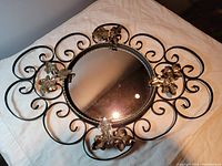 Top angled view of an oval convex wall mirror on a beige cloth showing the wrought iron frame with curling designs and four gold-coloured floral decorations around the glass.