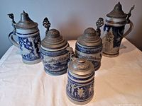 Five blue and gray stoneware beer steins with hinged pewter lids shown together from a front angle.