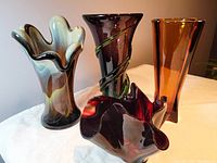 Four assorted handblown glass vases displayed on a table against a wall: purple and green vine vase, marbleized scalloped vase, amber Art Deco vase, and red slump vase.