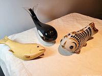 Three marine life figurines displayed on a white cloth background: glass orca, ceramic fish, and carved wooden fish.