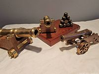Photo showing three brass and wood model cannons with moveable wheels and a wooden base with brass cannonballs in a triangular stack.