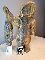 Front view of Inuit figure carved from fossilized whale bone, holding fish in left arm. Shows overall shape, coloration and tag.