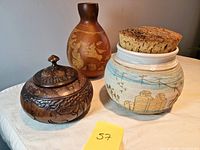 Image showing all three containers together on a table: wooden bowl with detailed carving and knob lid, lighter ceramic pot with cork lid, and tall calabash with animal carvings.