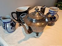 Photo showing silver-plated teapot with hinged lid alongside three small jugs on a white surface.