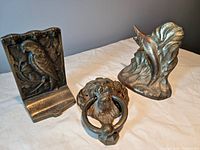 Three brass items displayed: lion head door knocker, parrot bookend, and marlin bookend shown on a cloth surface.