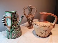 Three handmade international ceramic jugs: green and black painted jug with long spout and three handles, amphora-style jug decorated with tiny shells, and terra cotta jug with hand-painted designs.