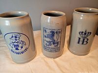 Photo of three large stoneware steins showing the front artwork featuring a cartoon Oktoberfest character, a lion emblem with Lowenbrau Muenchen text, and a crowned HB logo.