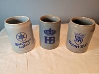 Front view of three half litre stoneware beer steins with brewery logos in blue.