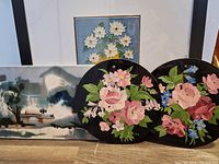Four pieces of ceramic and glass hand-painted wall art. Large framed floral tile, horizontal wooded landscape tile, and two black glass round floral paintings.