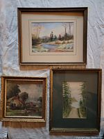 Three framed artworks laid out on white cloth: top frame contains river scene watercolour; bottom left frame contains country cottage print; bottom right frame contains wooded path watercolour.