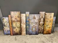 Front view of seven vertical wall art panels arranged side by side on carpet floor, showing different abstract and floral patterns in earth tone colors.