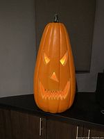 Front view of tall plastic pumpkin with lighted carved face, showing triangular eyes, carved nose, jagged mouth with teeth.