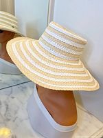 Side and angled views of the Nine West wide brim straw hat on mannequin head, showing beige and white striped pattern and small metallic logo detail on crown.