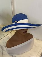 Hat shown on mannequin head with a curved wide brim, blue base color with white detailing and a white band with gold buckle.