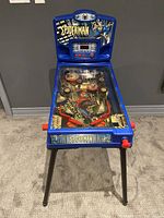 Front view of the blue Spider-Man pinball machine showing the playfield and legs on a carpeted floor against a gray wall.