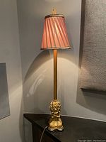 Full view of the lamp showing its tall gold-tone ornate base and pink pleated fabric shade, lamp switched on casting light on wall.