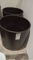 Two large black ceramic pots positioned side by side on carpeted floor, showing cylindrical shape and matte finish.