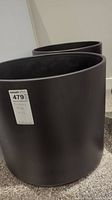 Two large black ceramic plant pots side by side on carpeted floor with white wall background. The pots are cylindrical with matte black finish. A price label is visible on one pot showing size 18x16.