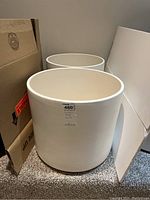 Photo showing two large white ceramic pots with cylindrical shape and smooth matte finish, positioned next to cardboard boxes.