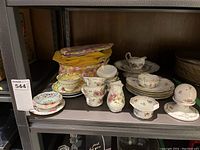 Assortment of cups, saucers, small bowls, creamer, sugar bowls, cake plate and decorative pierced bowl on metal shelving