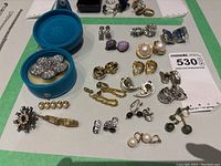 Wide view of assorted earrings and brooches displayed on a surface including a blue velvet box with jeweled earrings, pearl clip-ons, and various others in gold and silver tones.