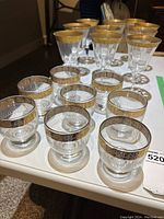 Overview of 8 short drinking glasses and 3 wine glasses with gold trim on a table.
