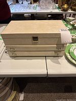 Closed beige jewellery box with gold-tone clasp on display table.