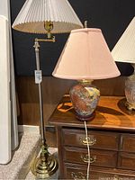 Photo of floor lamp and floral chinoiserie table lamp on wooden furniture against black background, showing tags and shades