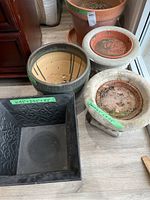 Photo showing multiple plant pots of various materials and sizes arranged on floor, including a large square black plastic planter and smaller round concrete and ceramic pots, some with dirt inside.