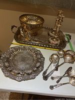 Displayed a collection of silver-plated serveware including bowls, trays, candlesticks, utensils, and salt and pepper shakers, arranged on a table