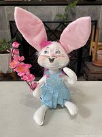 Front view of white Easter bunny doll with pink fabric ears, pink bow, blue overalls, holding pink flowers.