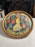 Photo of the front of the round wooden tray showing a detailed map of Finland with heraldic coats of arms and "SUOMI FINLAND" text in yellow on black background.
