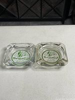 Top view of two square glass ashtrays with Spencer's Fish Grotto circular logos in green and white.