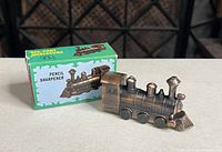 Photo showing the die-cast miniature train pencil sharpener with original box, highlighting the detailed antique finish and train shape.