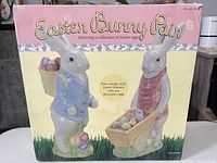 Box showing two Easter Bunny figurines carrying pastel Easter eggs, one with a basket and one with a wheelbarrow