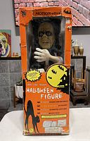 Front view of the vintage large ghoul Halloween figure in original orange box with spider and Halloween graphics, showing the figure inside with skeletal hands and face, box visibly damaged.