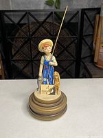 Front view of vintage Schmid Bros music box figurine showing boy in blue overalls holding a fishing pole and fish with dog beside him, mounted on a round gilt base.