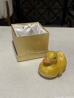 Yellow duck trinket box and gold-colored box with satin lining side view showing the glittery orange beak and the clasp.