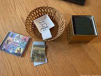 Photo of a small woven basket, a square ceramic dish with handwritten-style inscription, and a boxed CD titled 'The Sims Vintage Glamour Stuff'. Items are placed on a wooden surface.