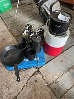 Photo of camping cookware and equipment on floor showing camp stove, cast iron skillet, kettle, mugs, thermoses, and red and white cooler.