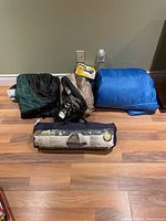 Sleeping bags and tent bag arranged on wooden floor; includes blue sleeping bag, green/black sleeping bag, and tent bag labeled '8 FT HEX DOME'