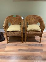 Front view of both wicker chairs with beige seat cushions placed on a wooden floor against a green wall.