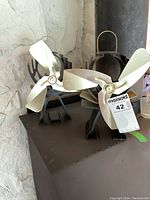 Two Ecofan stove fans shown on a dark surface in front of a white stone wall, each with three silver-colored blades and black metal bases, one with a MaxSold auction tag