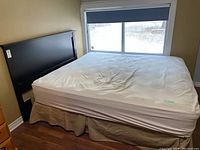 Bed set including black wooden headboard against wall, mattress and box spring with fitted sheet and beige fabric cover.
